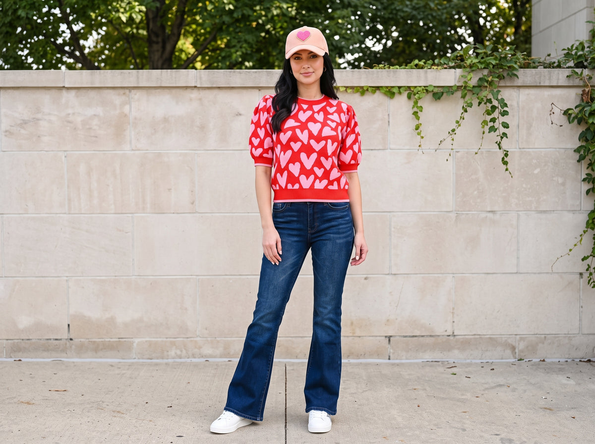 Woman in a pink heart baseball cap, red heart sweater, blue flare jeans and white sneakers