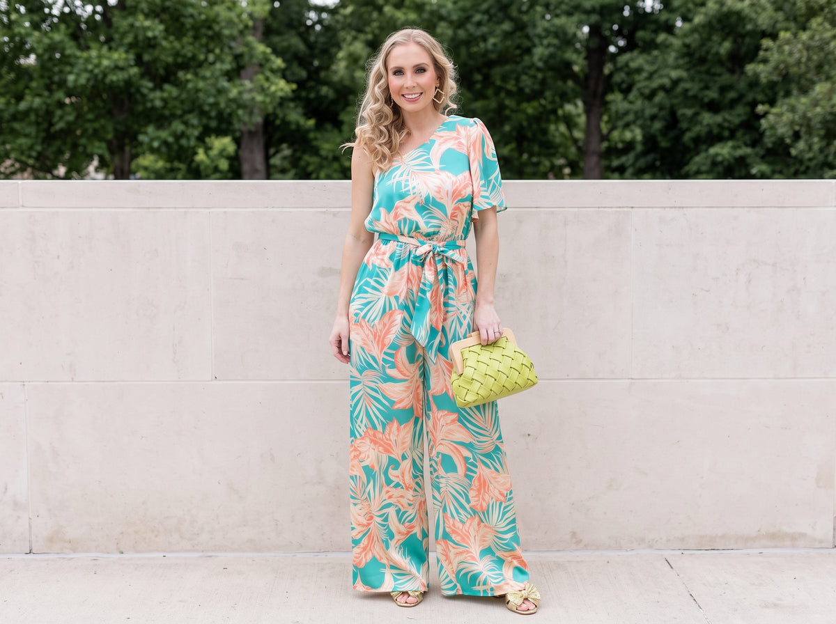 Woman in a turquoise and coral tropical print one-shoulder jumpsuit with gold bow sandals, a woven clutch and gold hoops