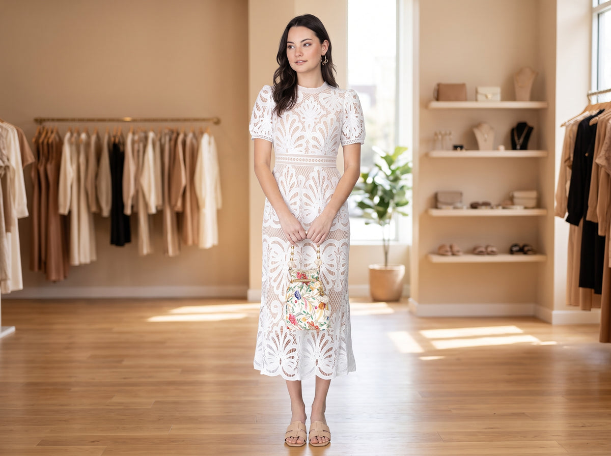 Woman in a white lace midi dress holding an ivory floral embroidered beaded-handle bag, with beige slide sandals and gold bow hoops