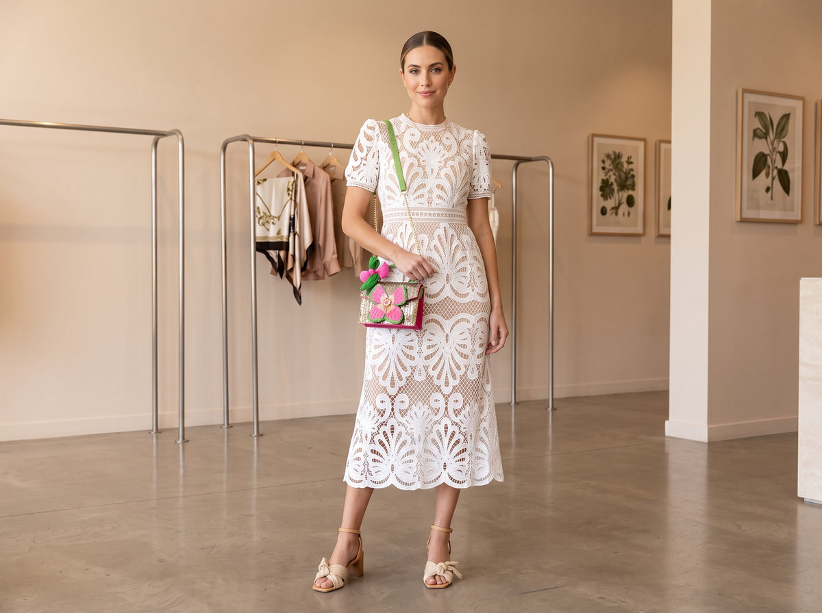 Woman in a white lace midi dress with a pink and green floral beaded bag on her shoulder and beige block heel sandals