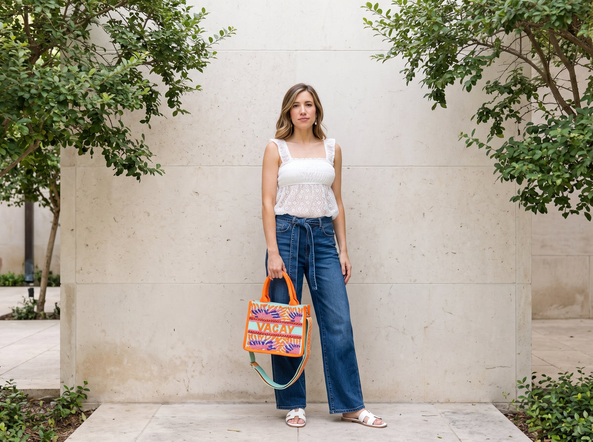 Woman in a white lace top and belted wide-leg jeans carrying an orange embroidered vacation handbag, with white slide sandals