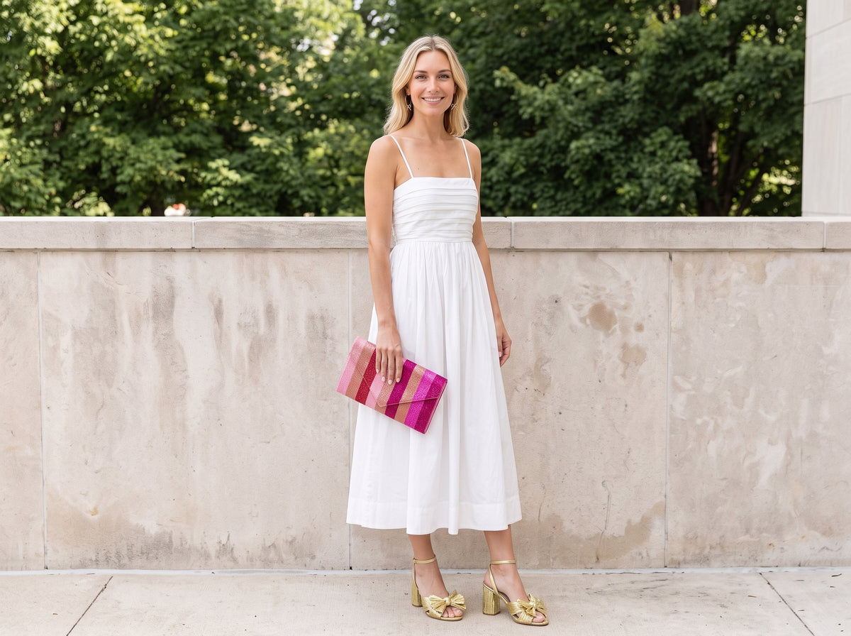 Woman in a white pleated midi dress and gold bow heeled sandals holding a pink striped beaded clutch