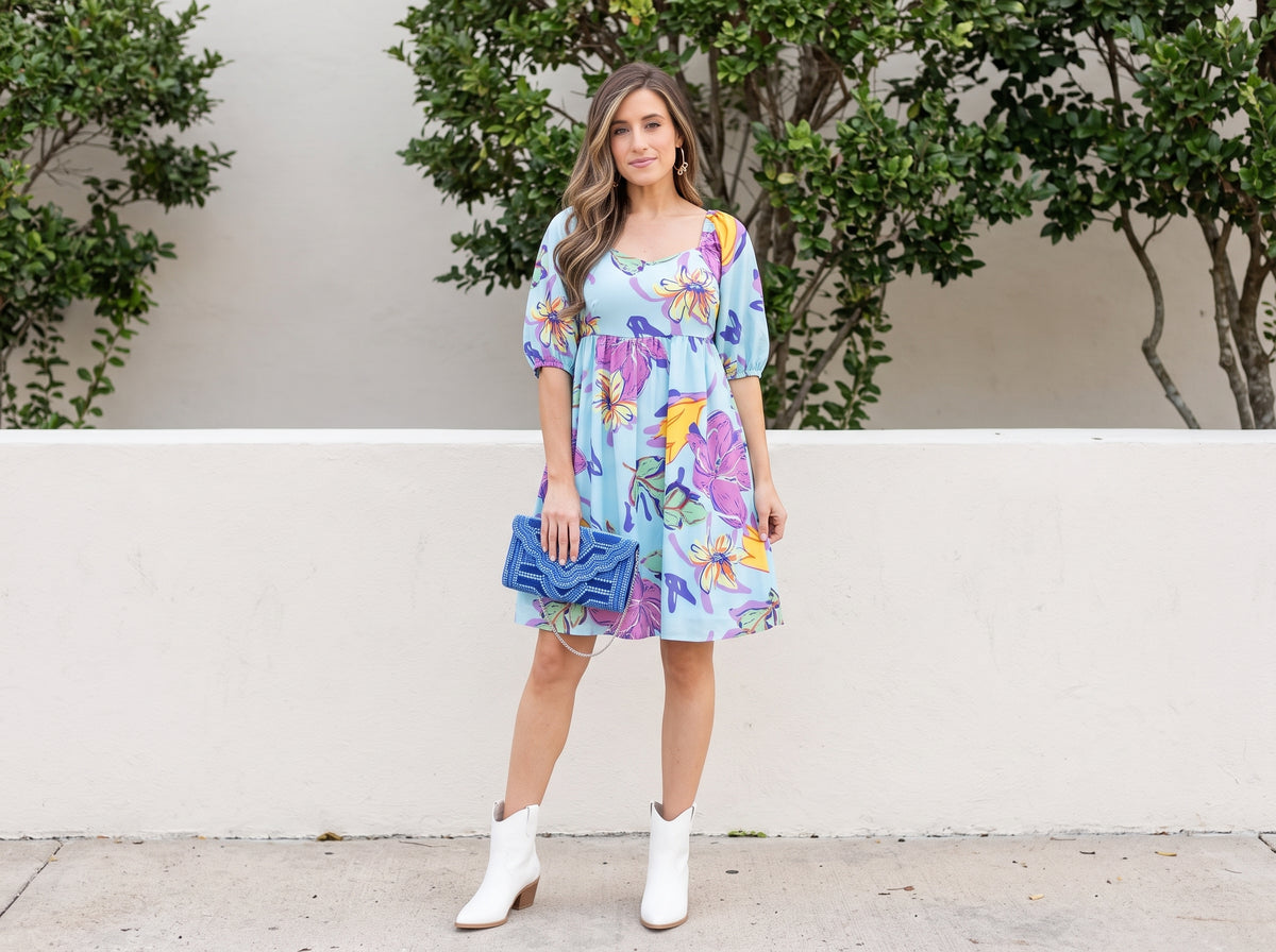 Woman in an aqua floral babydoll dress with white western booties, a blue clutch and gold bow hoop earrings