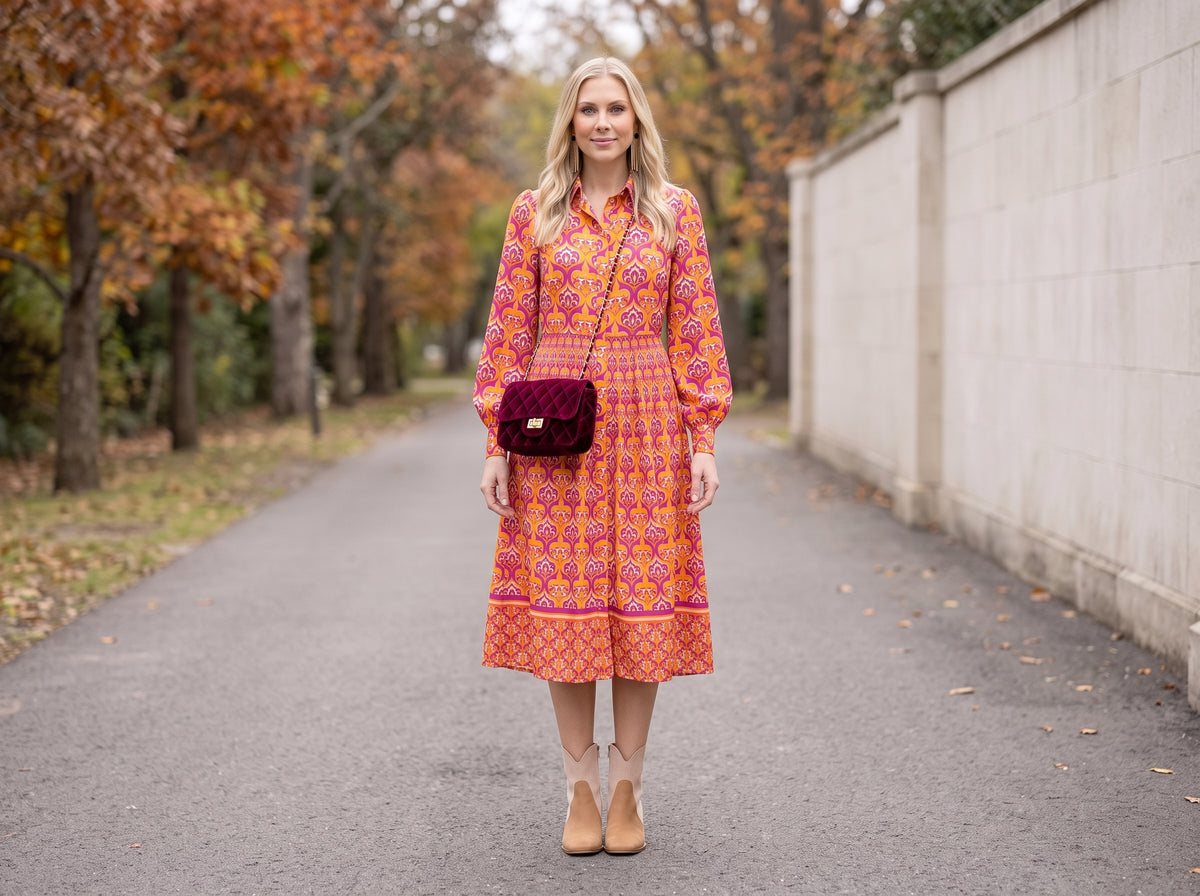 Woman in an orange and pink printed midi shirt dress with suede ankle booties, a burgundy velvet crossbody and gold fringe earrings