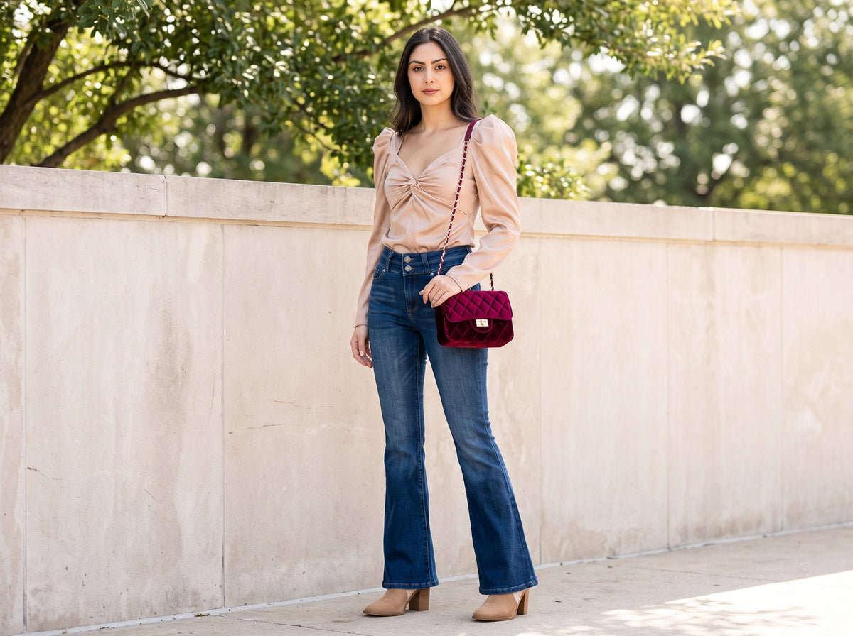 Woman in blue high-rise flare jeans with a champagne puff-sleeve top, suede booties and a burgundy velvet bag