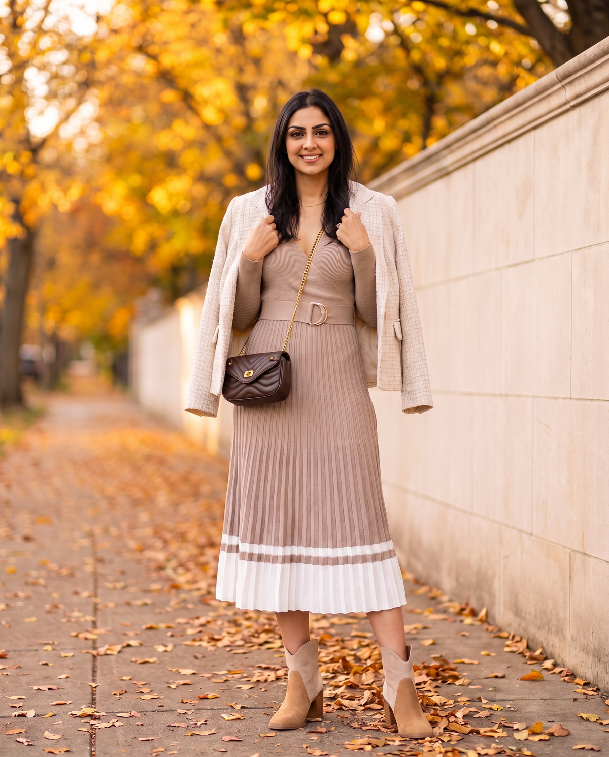 Woman on an autumn sidewalk in a beige pleated sweater dress with a belted waist and ivory hem, tri-tone suede ankle booties, a brown quilted crossbody bag and a cream tweed jacket over her shoulders