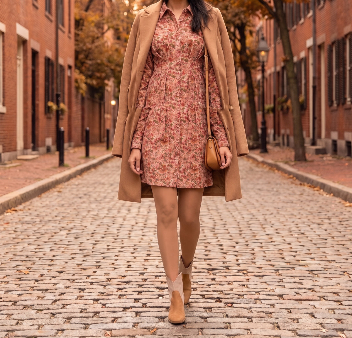 Woman walking down a cobbled autumn street in a floral corduroy mini shirt dress, a camel coat over her shoulders, a tan shoulder bag and tan suede ankle booties
