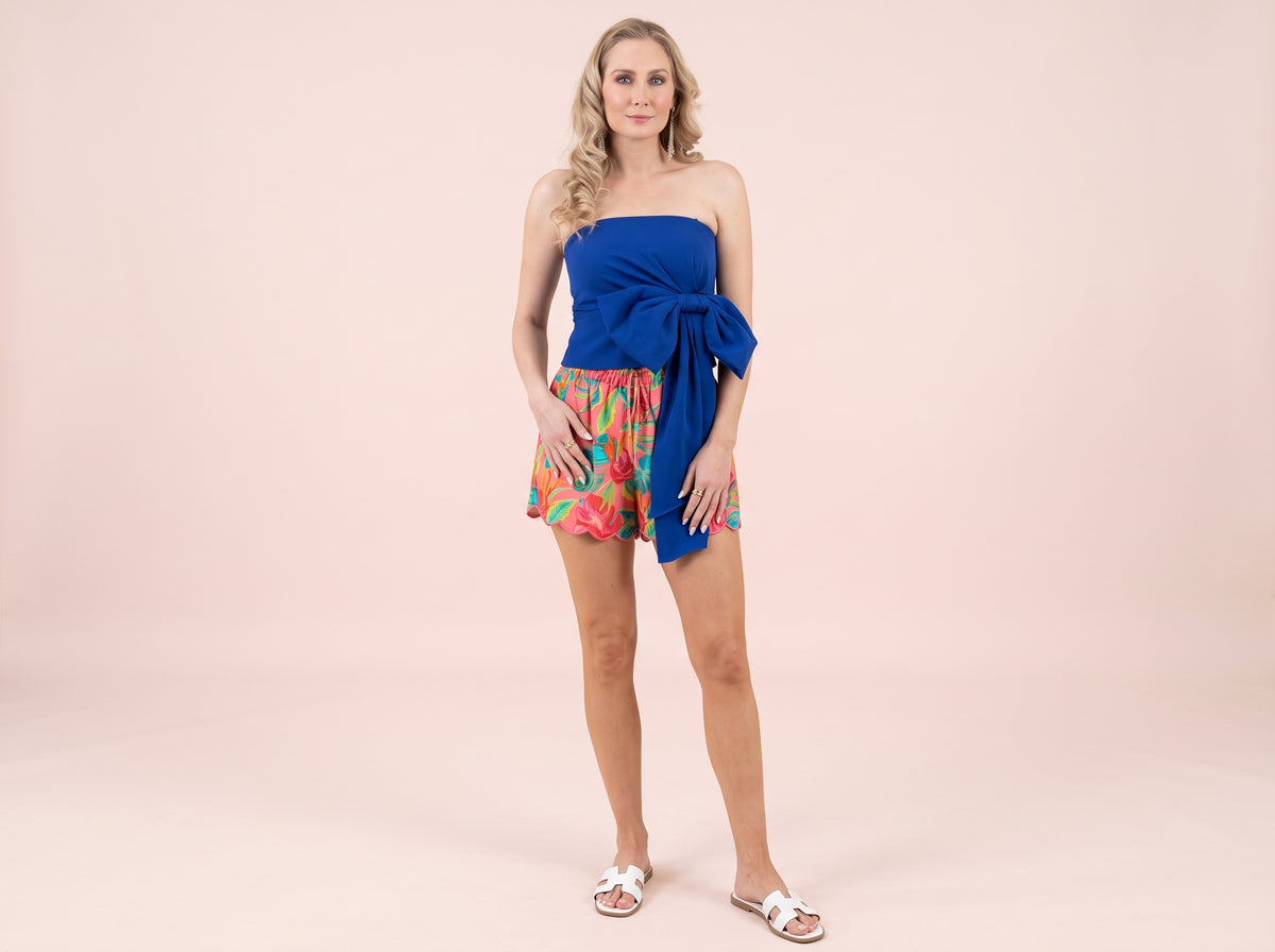 Woman wearing a gold double-band ring, a blue side-bow tube top, floral scalloped shorts and white slide sandals