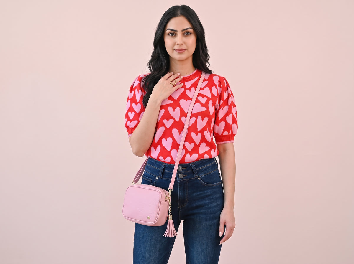 Woman wearing a gold open heart ring, a pink heart puff-sleeve sweater, high-waist flared jeans and a pink tassel crossbody