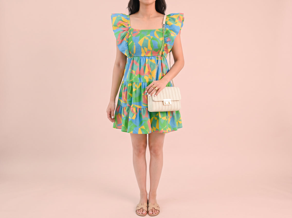 Woman wearing a gold open wrap ring, a green flutter-sleeve print mini dress, beige flat slide sandals and a cream braided crossbody