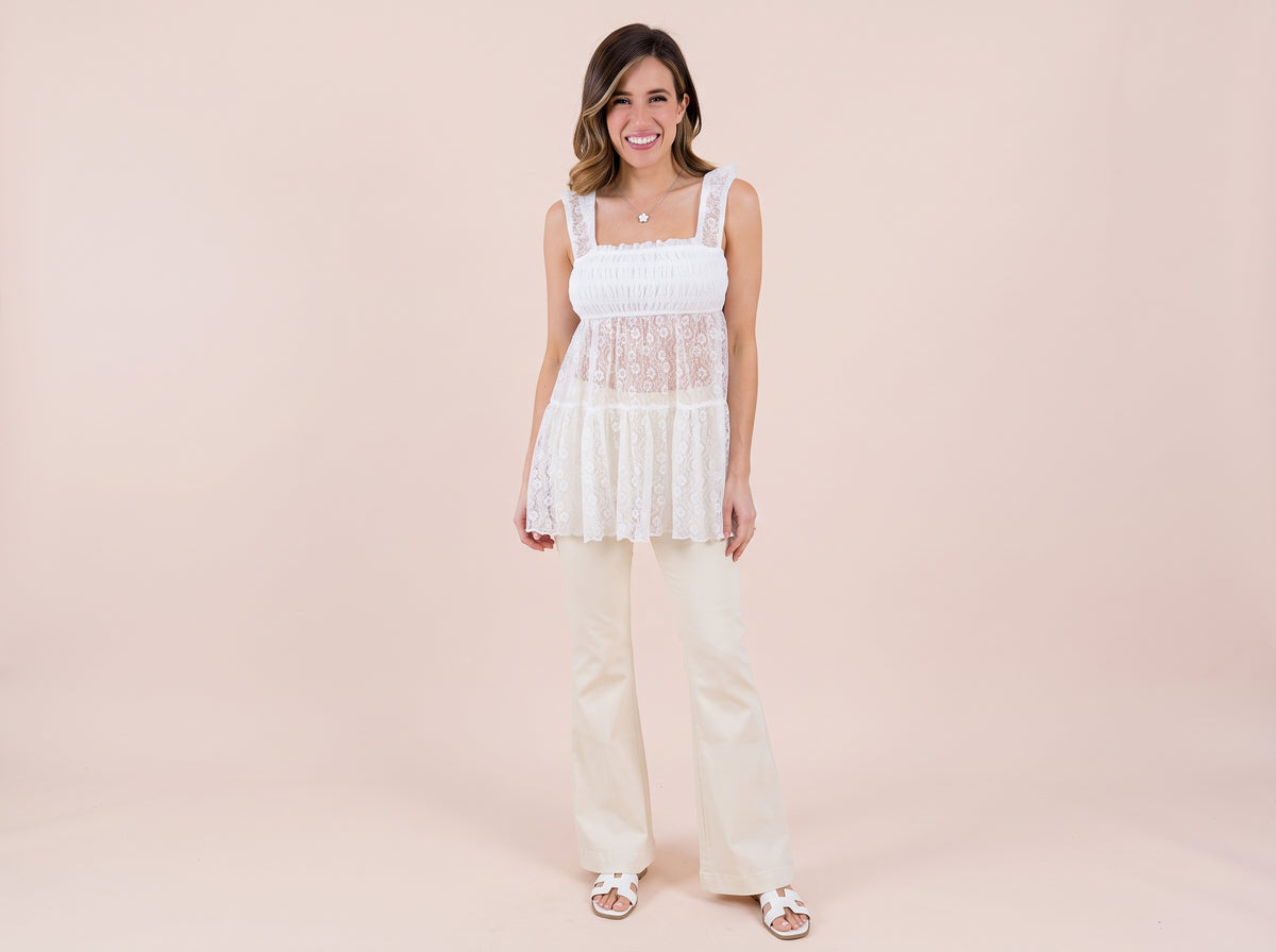 Woman wearing a silver flower pendant necklace, a white lace smocked top, cream flared jeans and white slide sandals