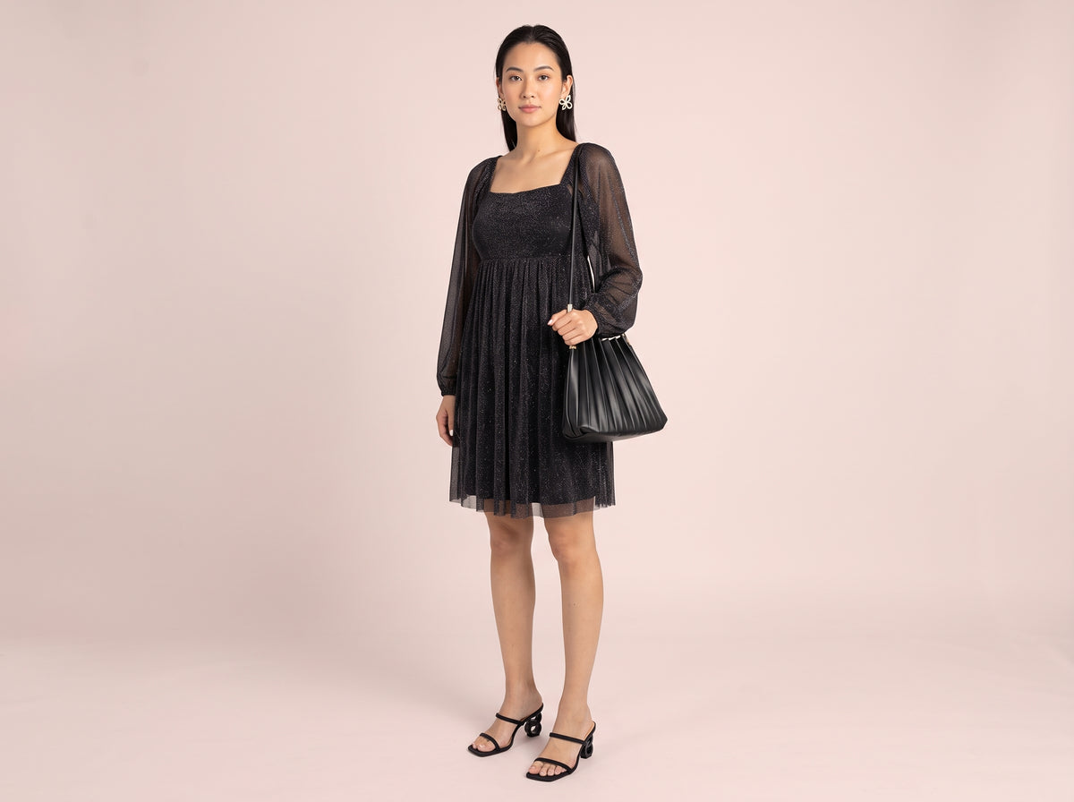 Woman wearing gold crystal bow stud earrings, a black glitter babydoll dress with sheer long sleeves and a black pleated shoulder bag