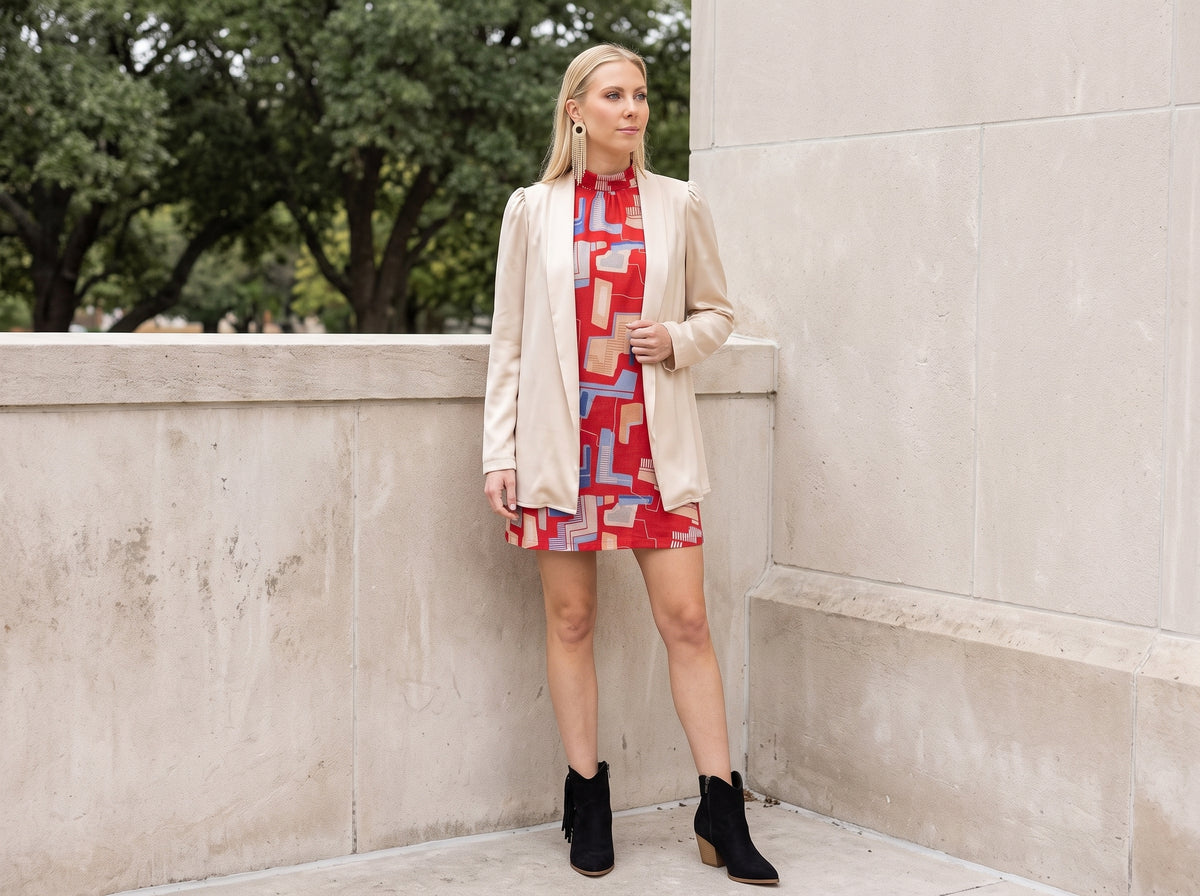 Woman wearing gold fringe earrings, a red geometric print shift dress, a beige blazer and black tassel ankle boots