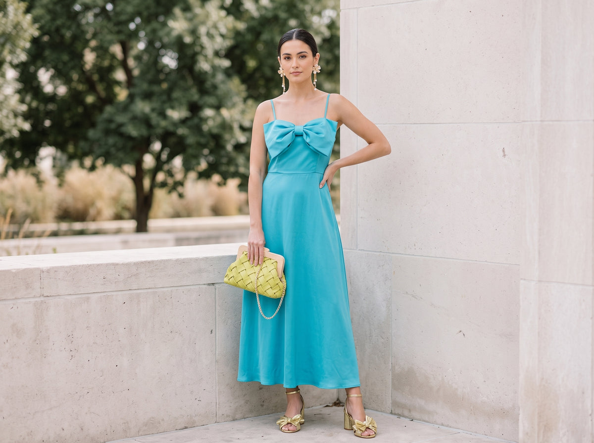 Woman wearing gold orchid flower earrings with pearl chains, a turquoise bow maxi dress, gold bow sandals and a woven clutch