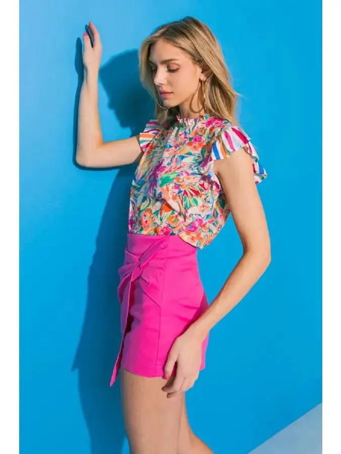 Model wearing a floral ruffle sleeve high neck top with vibrant colors against a blue background.