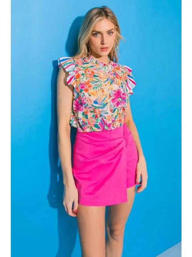 Model wearing a floral ruffle sleeve high neck top with vibrant colors against a blue background.